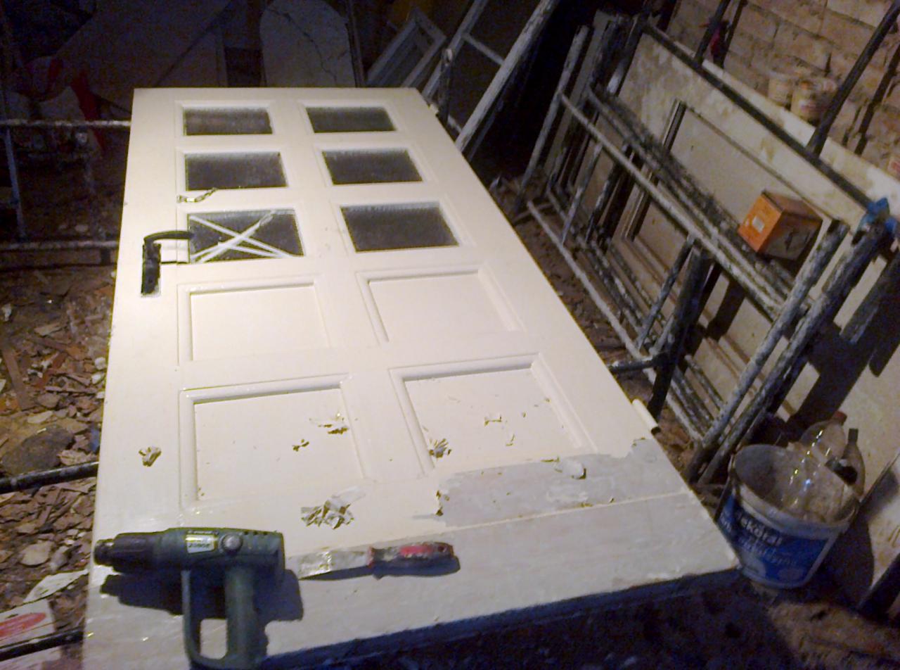 Białe drzwi z szybami, częściowo odrapane ze starej farby, leżące na boku z opalarką i szpachelką obok, w tle inne drzwi i drabina.