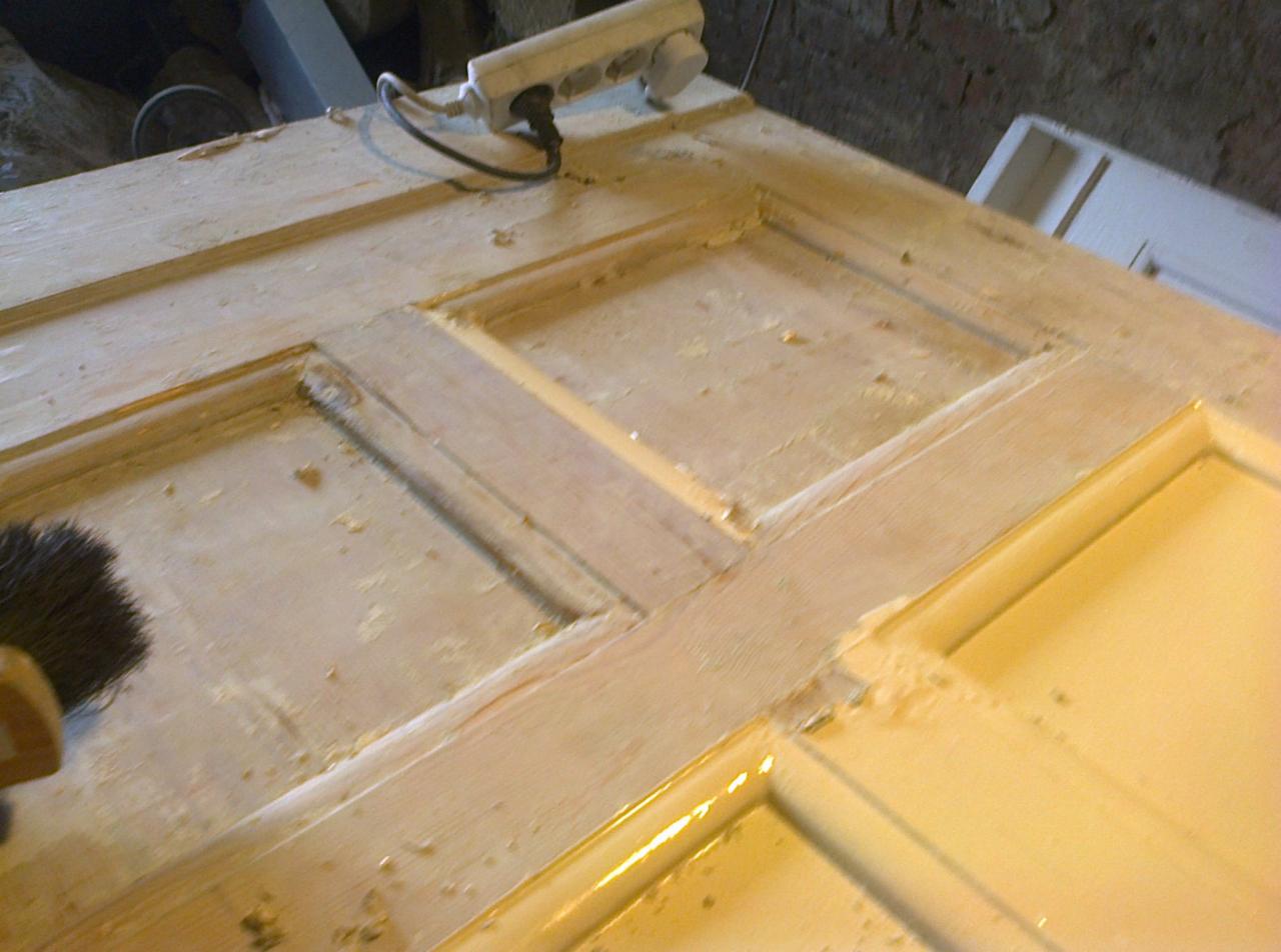 Drewniane drzwi z widocznymi śladami szlifowania i przygotowania do malowania, z brązowym pędzlem w lewym dolnym rogu i przedłużaczem elektrycznym w górnym rogu.