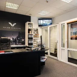 Wnętrze biura firmy Valor Windows z ekspozycją różnych typów okien i drzwi balkonowych, widoczne biurko z materiałami firmowymi i logo na ścianie.