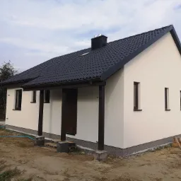 Jednopiętrowy budynek z białymi ścianami i ciemną dachówką, widoczne elementy drewniane, teren wokół domu częściowo zagospodarowany, niebieski wąż ogrodowy leżący na ziemi.