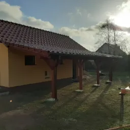 Żółty budynek gospodarczy z brązowym dachem i drewnianą wiatą, w tle widoczny drugi dom i drzewa.