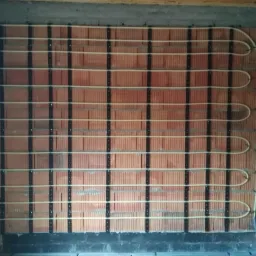 Rury ogrzewania podłogowego zamontowane na ścianie z cegły, widoczne czarne listwy mocujące i betonowy strop.