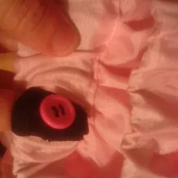 Różowa tkanina z falbanami, czarnym obszyciem i przyszytym różowym guzikiem z dwoma dziurkami, trzymana palcami.