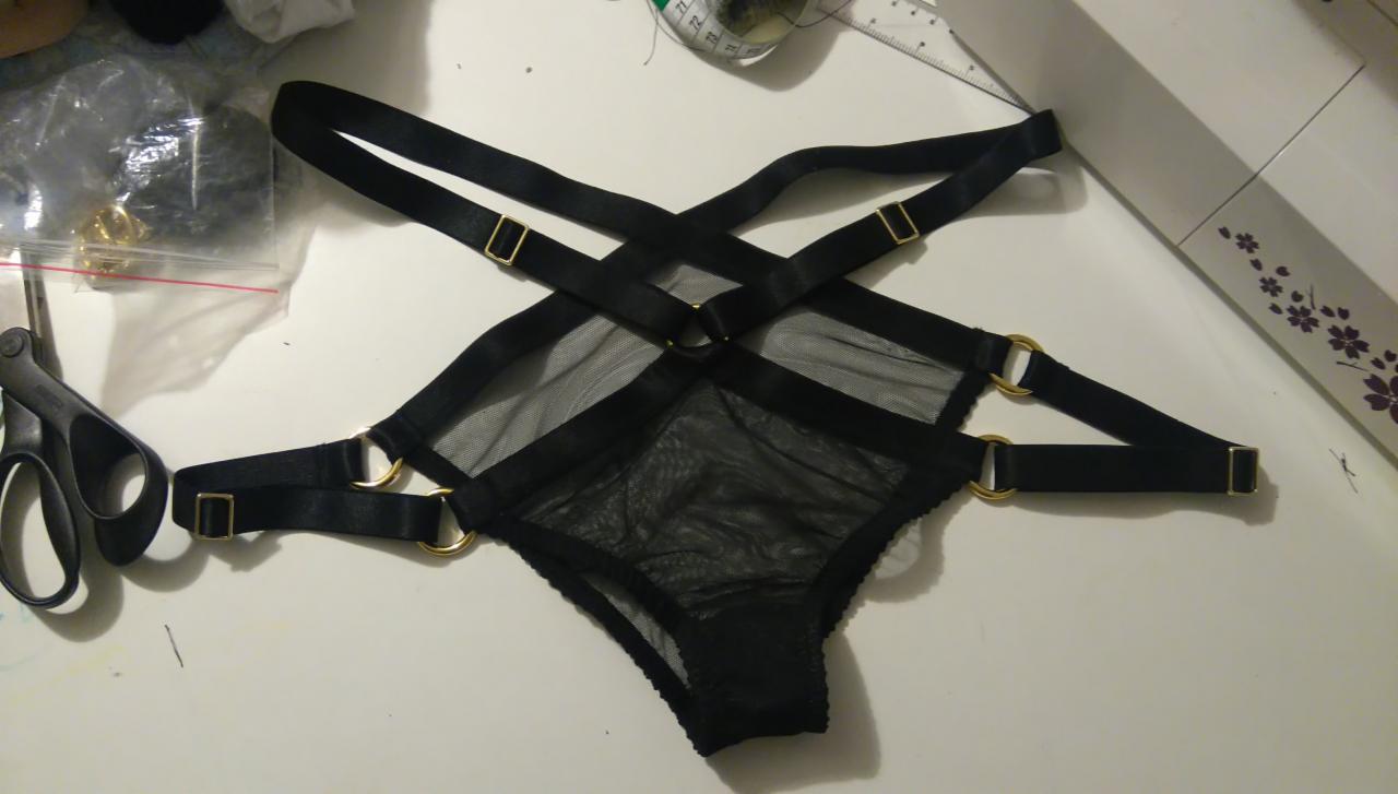 Czarne majtki z siateczki i paskami, z metalowymi kółkami i sprzączkami, leżące na stole obok nożyczek, miarki i maszyny do szycia.
