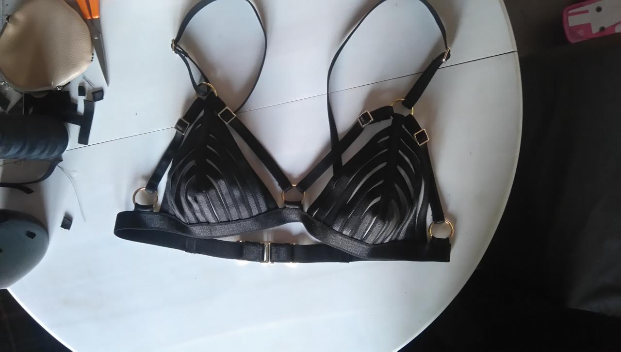 Czarny, ozdobny biustonosz z paskami i złotymi elementami leżący na białym stole, obok widoczne narzędzia krawieckie.