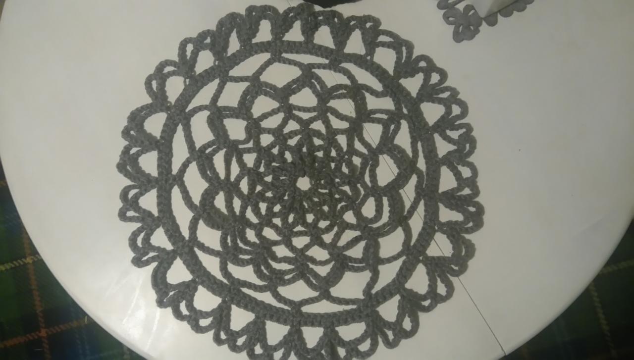 Szara, okrągła serwetka wykonana na szydełku, z ażurowym wzorem kwiatowym, leży na białym stole. W tle widoczny fragment zielonej tkaniny w kratę.