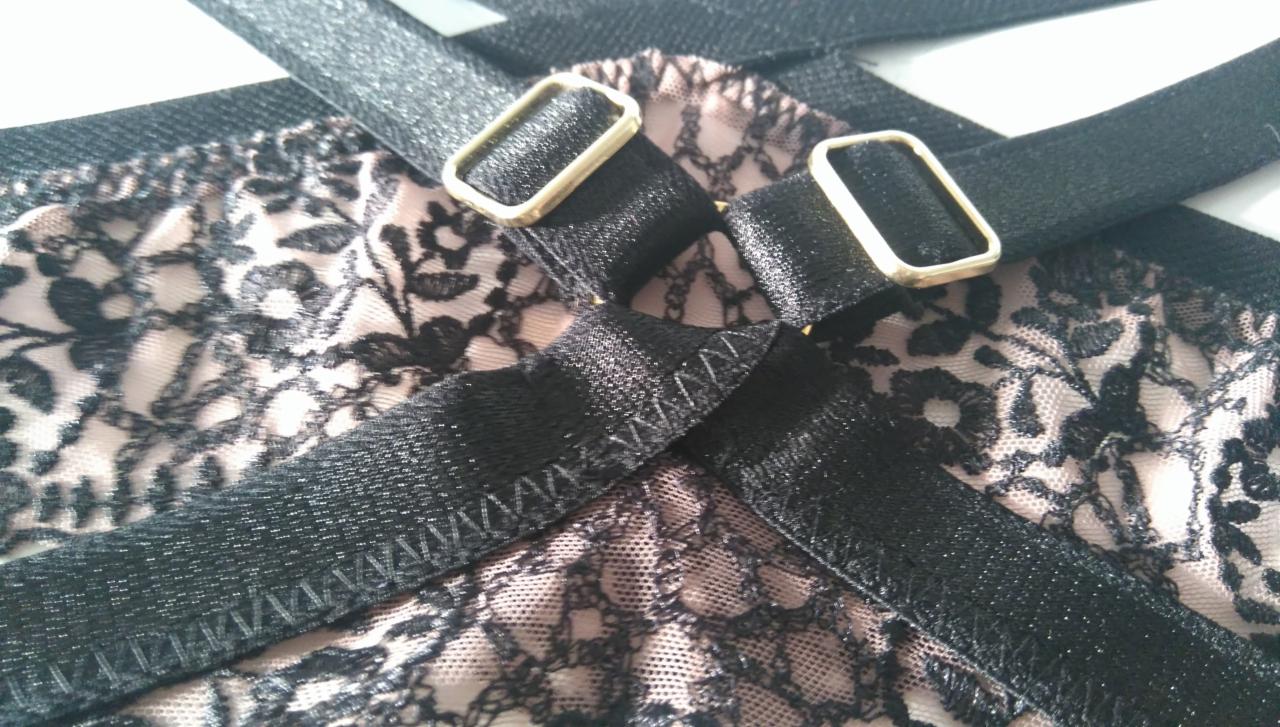 Fragment damskiej bielizny: czarna koronka na beżowym tle z regulowanymi, czarnymi ramiączkami z metalowymi sprzączkami, widoczne szwy.