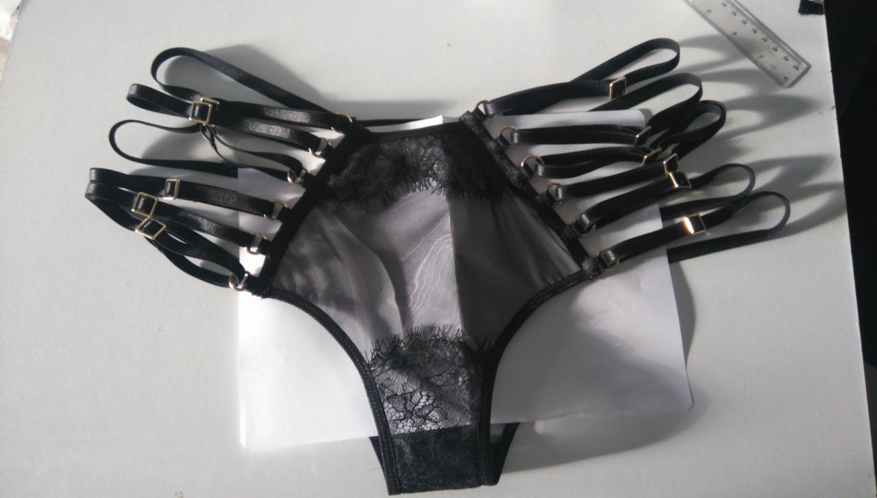 Czarne, koronkowe majtki typu stringi z paskami i złotymi sprzączkami, leżące na białym tle, obok linijki.