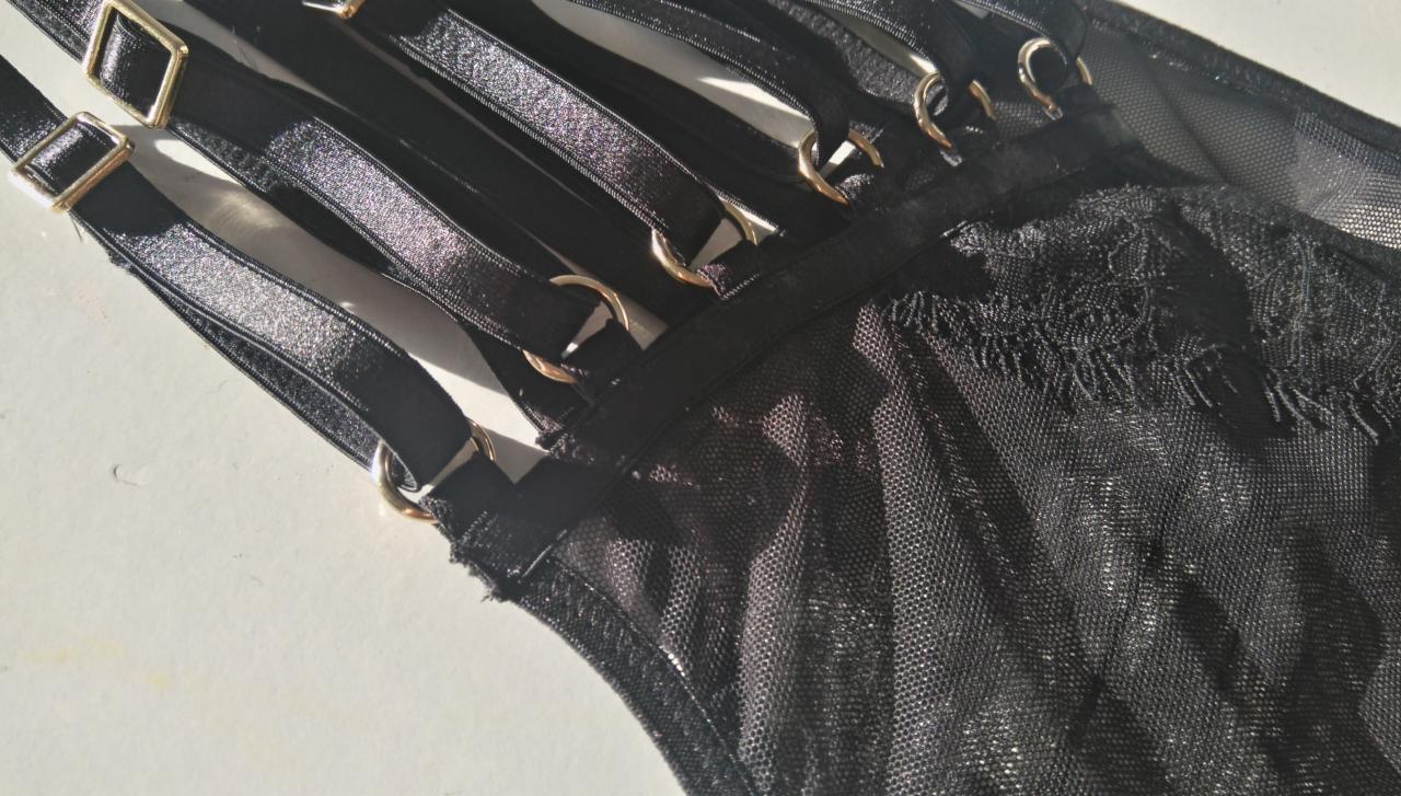 Fragment czarnej, koronkowej bielizny z ozdobnymi, regulowanymi ramiączkami i metalowymi kółkami, leżącej na jasnym tle.