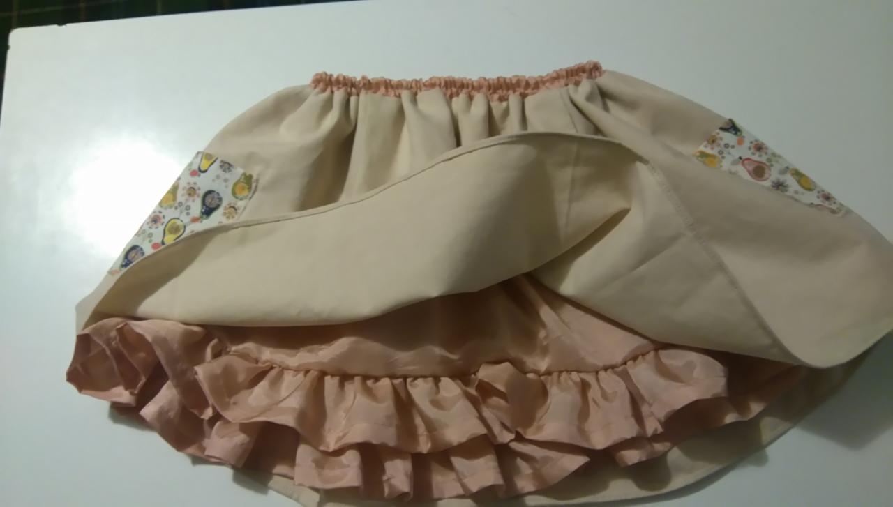 Beżowa spódniczka z falbanką i kieszeniami z motywem owocowym, widoczne marszczenie w pasie.