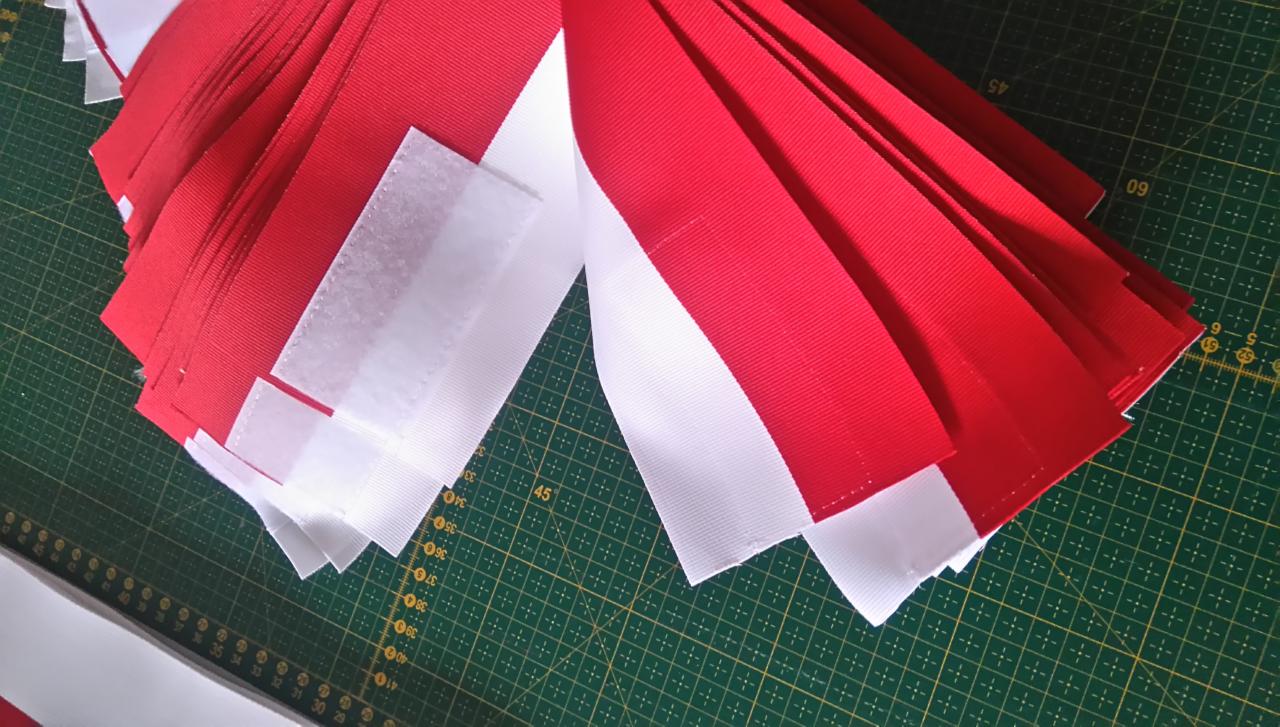 Przygotowane paski czerwonej i białej rypsowej wstążki z rzepem, ułożone warstwowo na zielonej macie krawieckiej z podziałką.