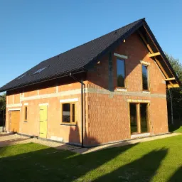 Niedokończony dom jednorodzinny z czerwonej cegły ceramicznej, z czarnym dachem i zamontowanymi oknami, widoczne drewniane elementy konstrukcji dachu, położony na działce z trawnikiem i drzewami.