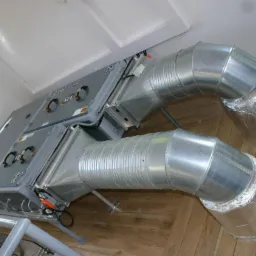 Szara centrala wentylacyjna z dwoma kanałami wentylacyjnymi z blachy falistej, izolowanymi folią aluminiową na końcach, zamontowana w pomieszczeniu z drewnianą podłogą.