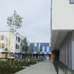 Nowoczesny budynek w Warszawie z kolorową elewacją w odcieniach niebieskiego, żółtego i białego, widoczny z perspektywy chodnika z kostki brukowej i zielenią.