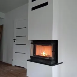 Nowoczesny kominek z białym obudowaniem i czarnymi akcentami, płomienie widoczne przez szklaną szybę, drewno obok, drzwi w tle, podłoga wyłożona panelami imitującymi drewno.