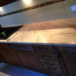 Beżowy blat kuchenny z wzorem marmuru, wbudowany czarny zlew, dolne szafki z ciemnego drewna i srebrnymi uchwytami, białe płytki z dekoracyjnym paskiem mozaiki podświetlone punktowym światłem.