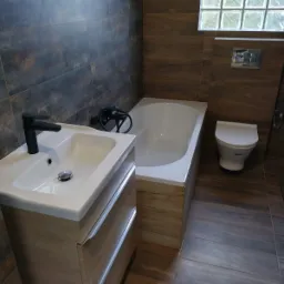 Nowoczesna łazienka z brązowymi płytkami imitującymi drewno, białą armaturą i czarną baterią umywalkową.