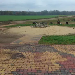 Przygotowanie terenu pod budowę: wyrównany teren piaskiem, hałda kamieni, żółty wąż, częściowo ułożona kostka brukowa, widok na pole i las w tle.