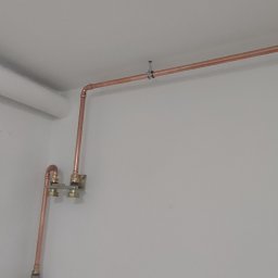 Miedziana instalacja wodna z zaworami, zamontowana przy suficie w pomieszczeniu o białych ścianach. Widoczne mocowania rur i fragment instalacji wentylacyjnej.