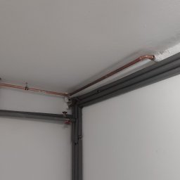 Miedziane rury CO pod sufitem, częściowo zaizolowane szarą otuliną. Widoczne zawory i mocowania. Białe ściany i sufit. Ujęcie z dołu, w rogu pomieszczenia.