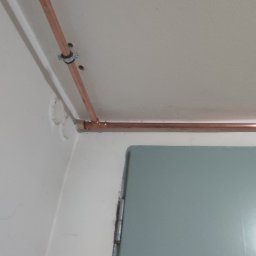 Miedziana instalacja gazowa przy suficie, zamocowana obejmą, widoczne łączenie rur. Szare futryny okna i drzwi. Białe ściany i sufit. Ujęcie z dołu, blisko narożnika.