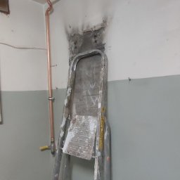 Miedziane rury instalacji CO prowadzone wzdłuż ściany i sufitu, drabina malarska oparta o ścianę. Widoczne ślady po kuciu w ścianie.