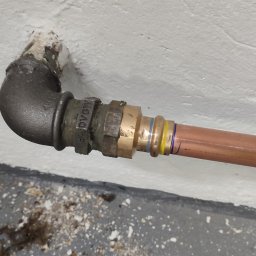 DRENIK INSTALACJE Tomasz Drenikowski - Miedziana rura połączona z żeliwnym kolankiem przy ścianie. Widoczne ślady uszczelniania i oznaczenia na rurze. Instalacja po montażu, gotowa do użytku.