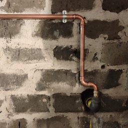 DRENIK INSTALACJE Tomasz Drenikowski - Miedziana instalacja rurowa, zamocowana do surowej ściany z cegły, z widocznymi połączeniami lutowanymi i uchwytem mocującym. Widoczny fragment zaworu.