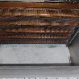 DRENIK INSTALACJE Tomasz Drenikowski - Miedziany wymiennik ciepła wewnątrz obudowy pieca gazowego, widoczne ślady użytkowania i osady. Ujęcie z góry, wnętrze pieca oświetlone.