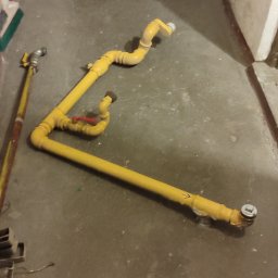DRENIK INSTALACJE Tomasz Drenikowski - Żółte rury gazowe z zaworem na szarym betonowym podłożu, widoczne złączenia i elementy instalacji. Fragment ściany i drzwi w tle.
