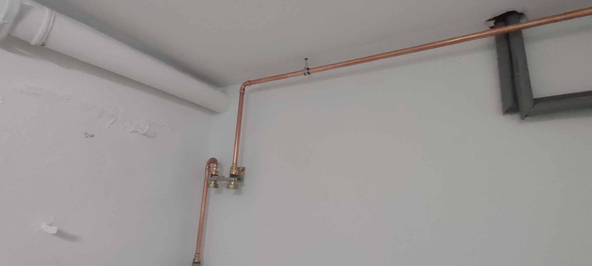 Miedziana instalacja wodna z zaworami, zamontowana przy suficie w pomieszczeniu o białych ścianach. Widoczne mocowania rur i fragment instalacji wentylacyjnej.