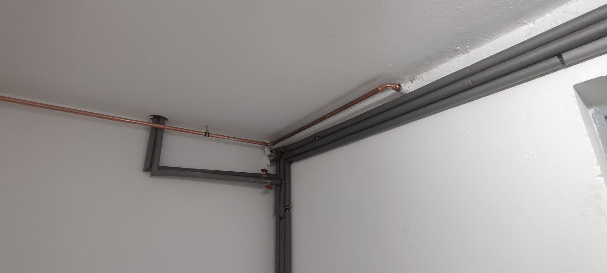 Miedziane rury CO pod sufitem, częściowo zaizolowane szarą otuliną. Widoczne zawory i mocowania. Białe ściany i sufit. Ujęcie z dołu, w rogu pomieszczenia.