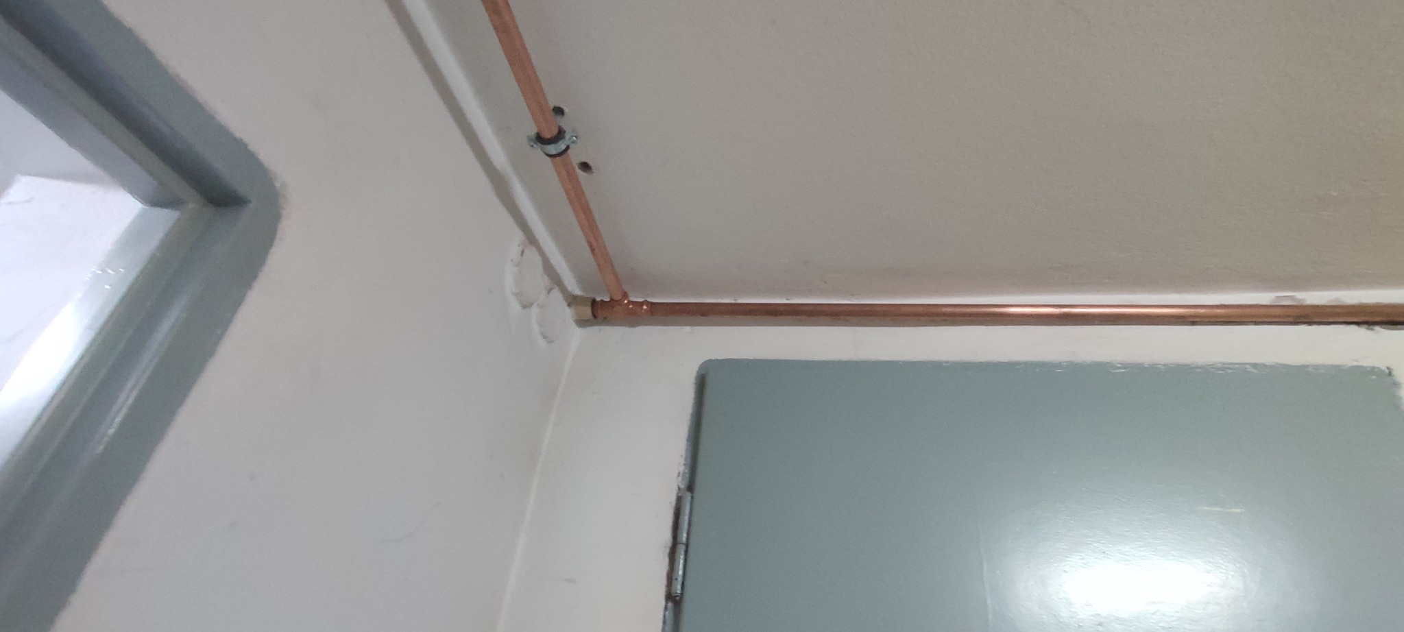 Miedziana instalacja gazowa przy suficie, zamocowana obejmą, widoczne łączenie rur. Szare futryny okna i drzwi. Białe ściany i sufit. Ujęcie z dołu, blisko narożnika.