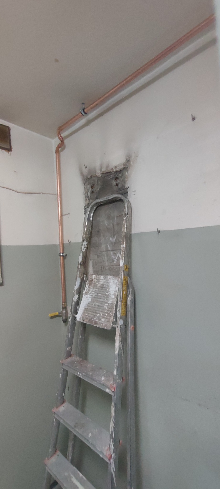 Miedziane rury instalacji CO prowadzone wzdłuż ściany i sufitu, drabina malarska oparta o ścianę. Widoczne ślady po kuciu w ścianie.