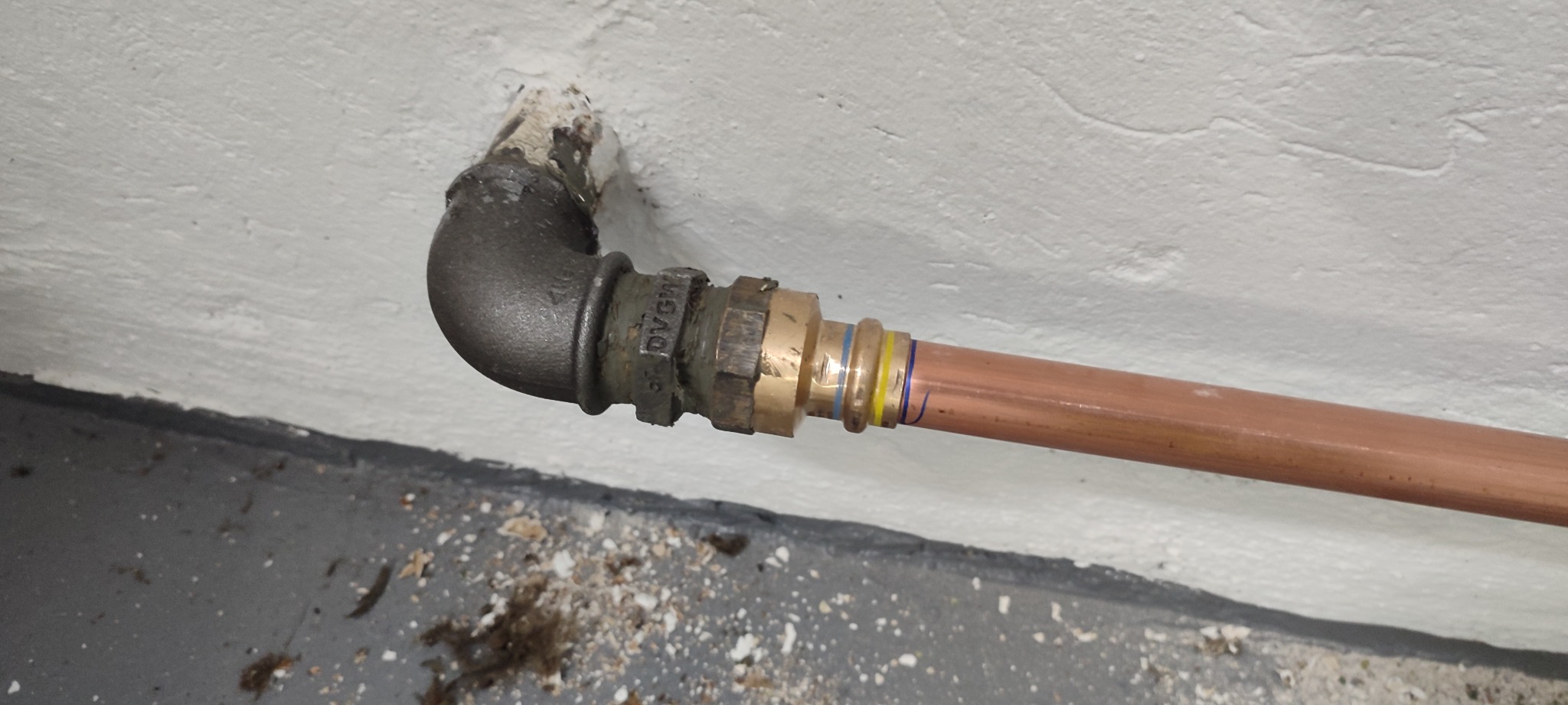 Miedziana rura połączona z żeliwnym kolankiem przy ścianie. Widoczne ślady uszczelniania i oznaczenia na rurze. Instalacja po montażu, gotowa do użytku.