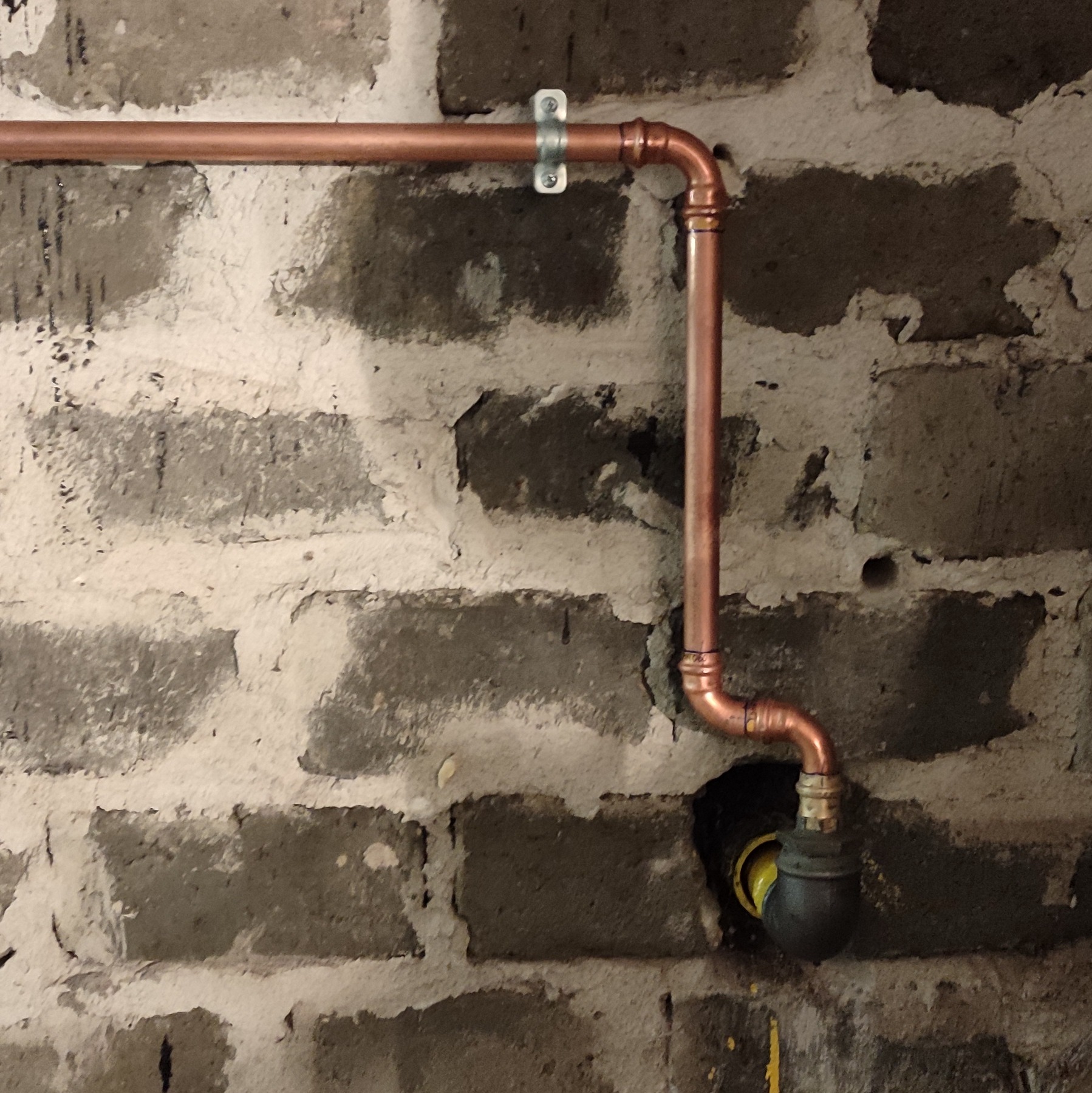 Miedziana instalacja rurowa, zamocowana do surowej ściany z cegły, z widocznymi połączeniami lutowanymi i uchwytem mocującym. Widoczny fragment zaworu.