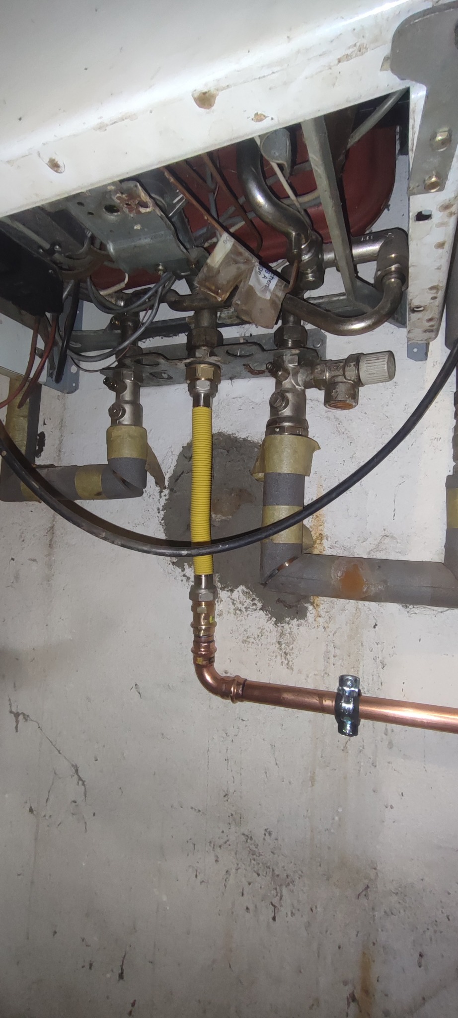 Instalacja gazowa z żółtym przewodem elastycznym i miedzianą rurą. Widoczne elementy pieca gazowego i ściana z szarą farbą. Detal przyłącza gazowego.