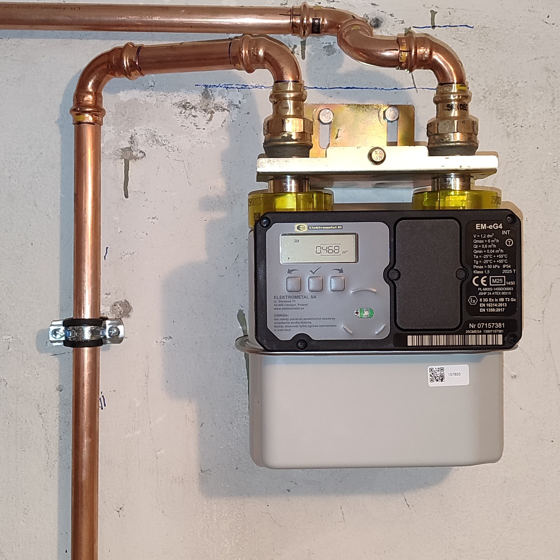 Zamontowany gazomierz Elektrometal z miedzianą instalacją, widoczne złącza i mocowania. Szara ściana w tle. Precyzyjny montaż instalacji gazowej.