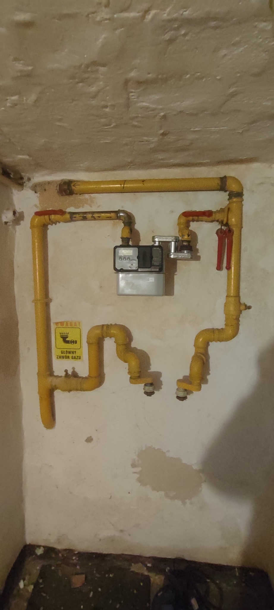 Żółta instalacja gazowa z licznikiem i zaworami na tle ściany z widocznymi śladami użytkowania. Na rurach naklejka ostrzegawcza.
