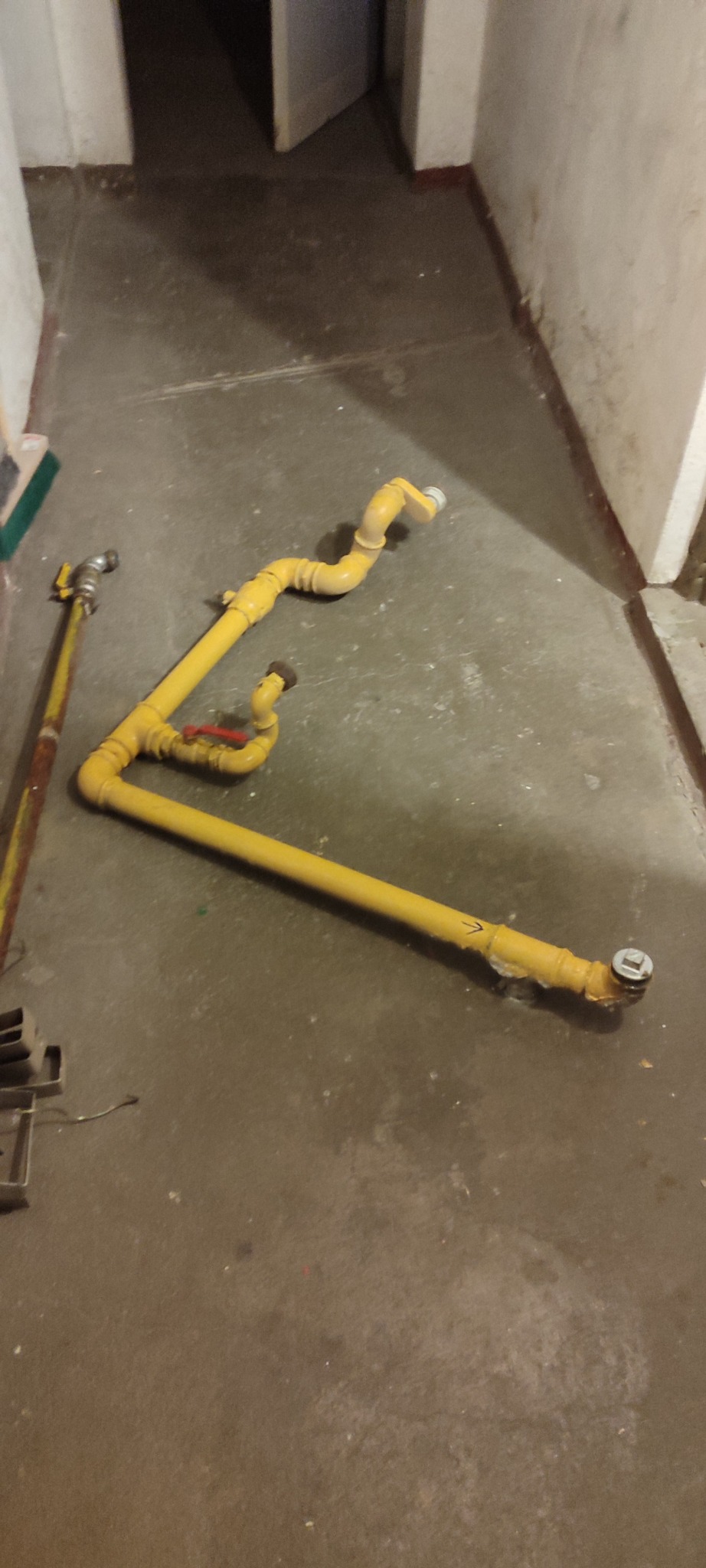Żółte rury gazowe z zaworem na szarym betonowym podłożu, widoczne złączenia i elementy instalacji. Fragment ściany i drzwi w tle.