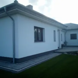 Nowoczesny, parterowy dom z białymi ścianami i szarą podmurówką, widoczny z zewnątrz, z równo przystrzyżonym trawnikiem i brukowanym chodnikiem.