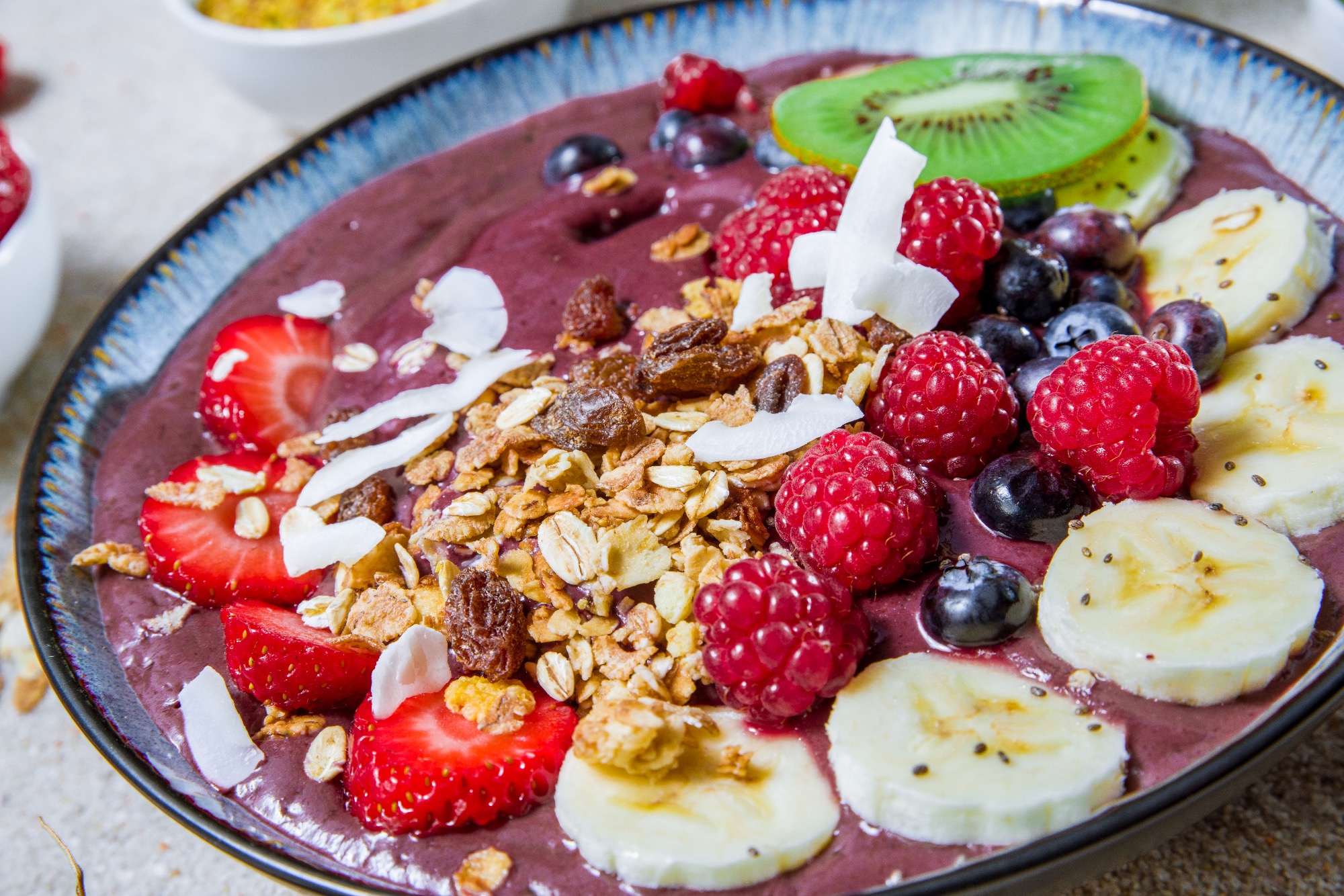 Acai bowl z truskawkami, malinami, borówkami, bananami, kiwi, płatkami kokosowymi i granolą, podana w ceramicznej misce z niebieską obwódką.