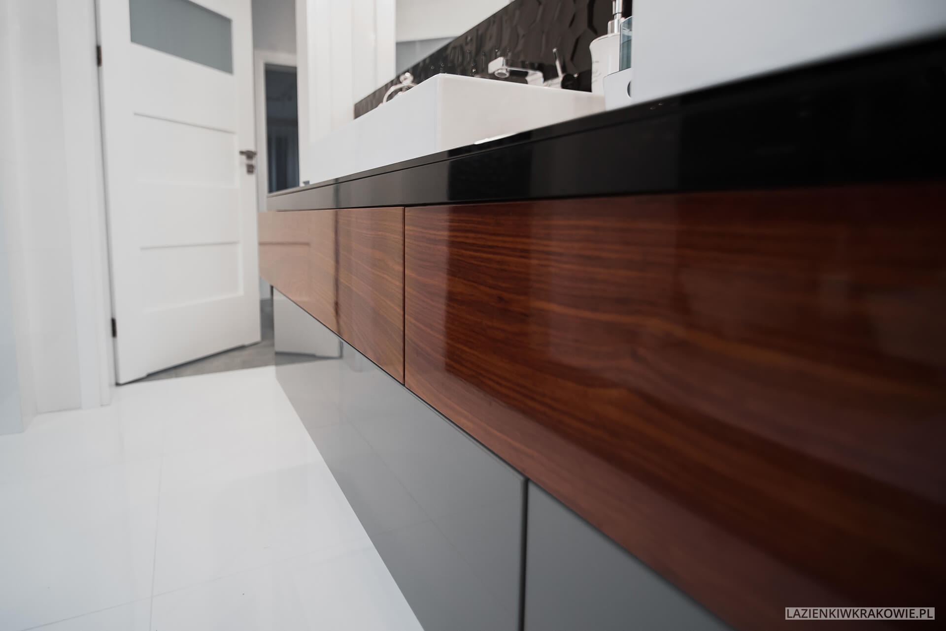 Nowoczesna łazienka: wisząca szafka pod umywalkę z frontami z ciemnego drewna o wyraźnym usłojeniu, szara podstawa, biała umywalka nablatowa i czarne płytki dekoracyjne na ścianie w tle, biała...