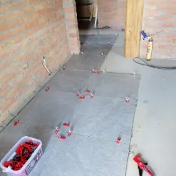 Układanie szarych płytek podłogowych z użyciem czerwonych klipsów poziomujących, widoczna ściana z cegły i puszka z pianką montażową.