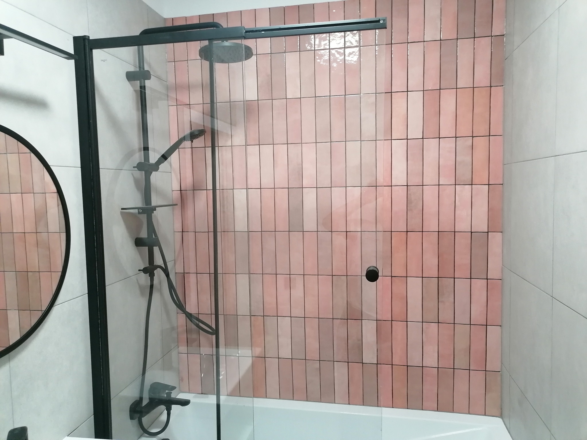 Nowoczesna kabina prysznicowa z czarnymi elementami i różowymi, błyszczącymi płytkami ułożonymi pionowo, widoczna okrągła rama lustra.