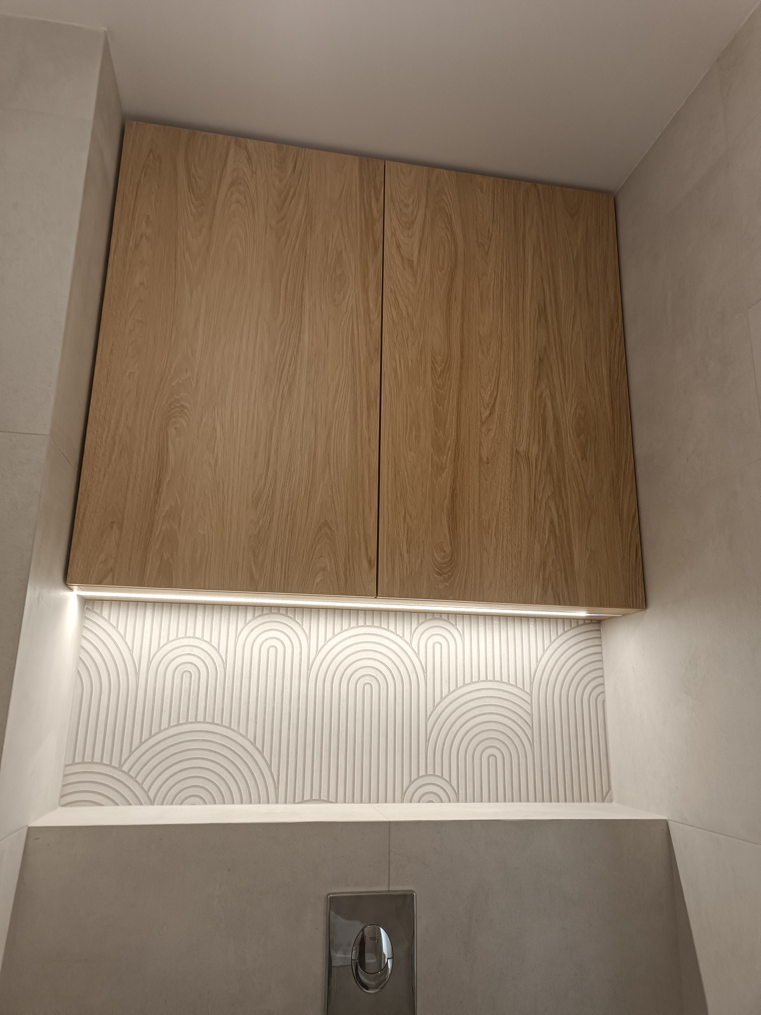 Drewniane szafki wiszące nad toaletą z podświetleniem LED i dekoracyjną mozaiką w łazience. Minimalistyczny design, ciepłe oświetlenie.