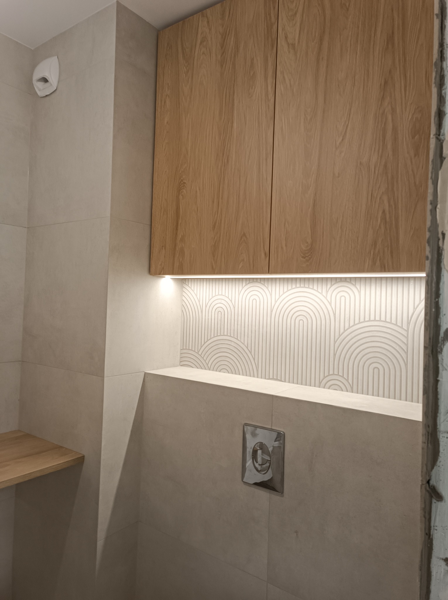 Wykończona łazienka z drewnianą szafką, podświetlaną półką i wzorzystą ścianą. Widoczny fragment surowej ściany w trakcie prac wykończeniowych.