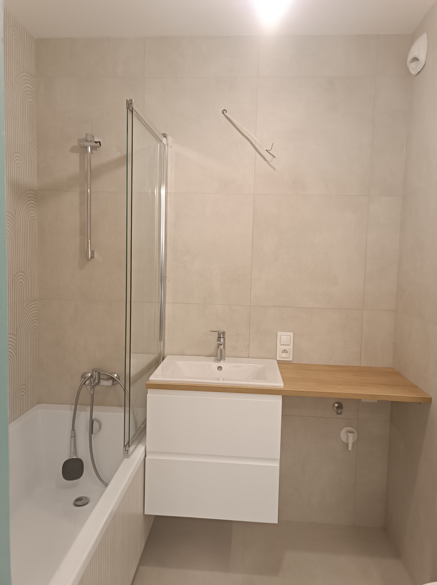 Jasna łazienka po remoncie: biała umywalka z drewnianym blatem, wanna z parawanem, beżowe płytki. Widoczne przyłącza elektryczne i hydrauliczne.