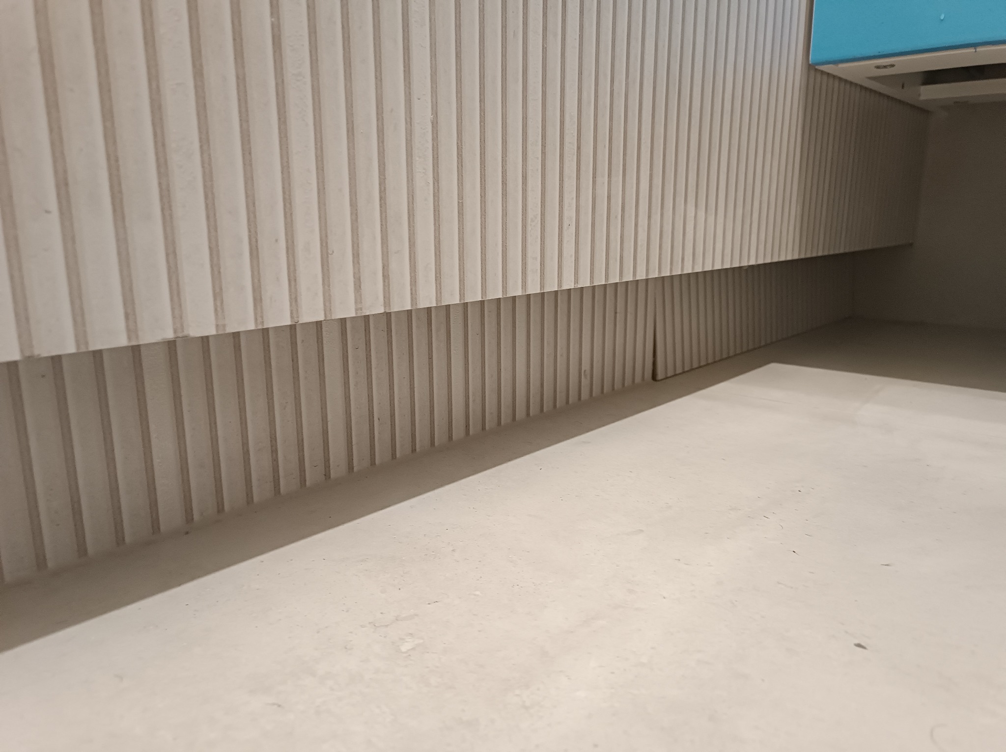 Biała, ryflowana zabudowa meblowa pod umywalką z turkusowym blatem, widoczna z niskiej perspektywy, podkreślająca minimalistyczny design i gładką powierzchnię podłogi.