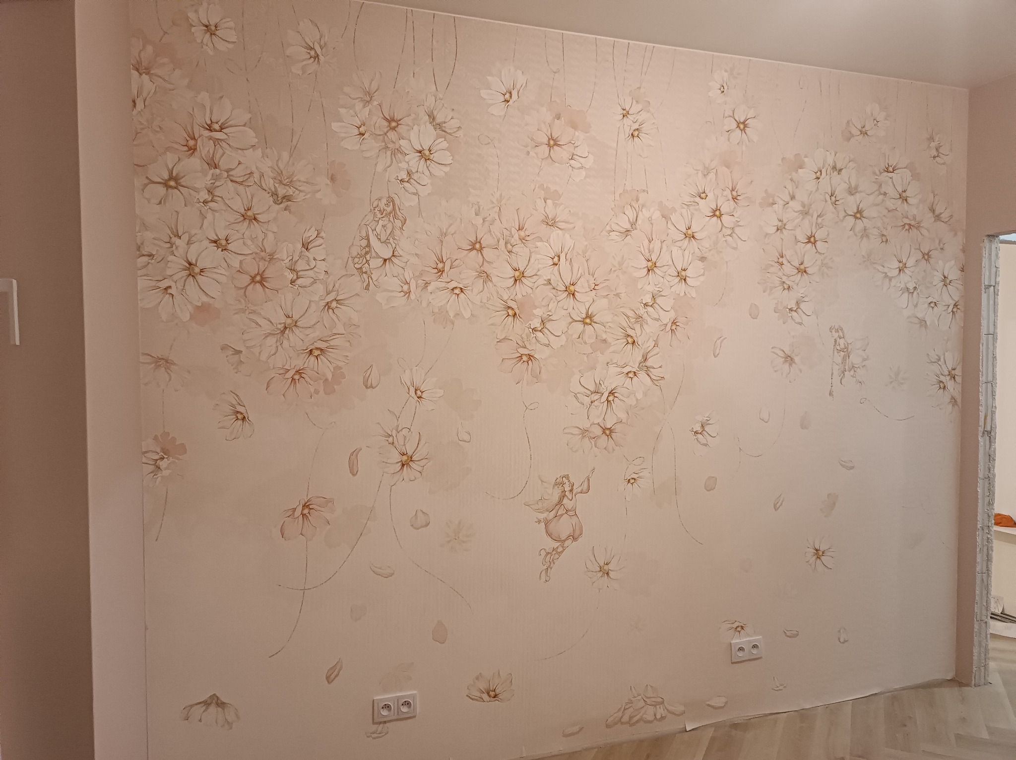 Ściana z tapetą w kwiaty i postać wróżki, gniazdka elektryczne na dole. Wykończenie wnętrza w pastelowych barwach, jasne drewniane panele podłogowe.